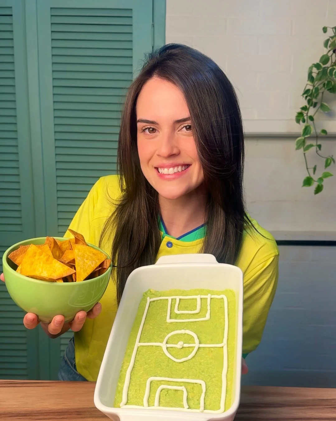 Elize sorrindo e mostrando uma receita em uma travessa com desenho de campo de futebol